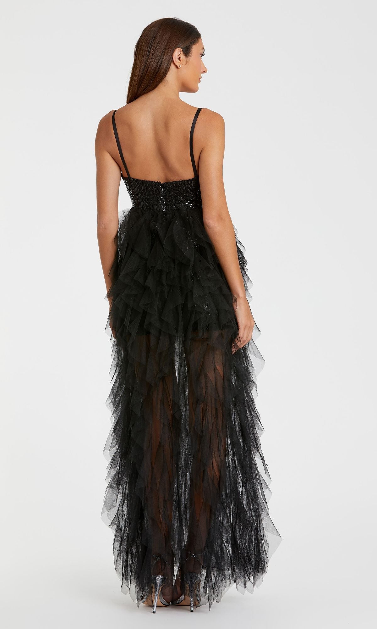 A woman models the Mac Duggal 27119 Overskirt Mini Sequin Prom Dress, featuring a sequined bodice, thin straps, and a sheer layered tulle skirt. She pairs it with silver heels against a plain white background.
